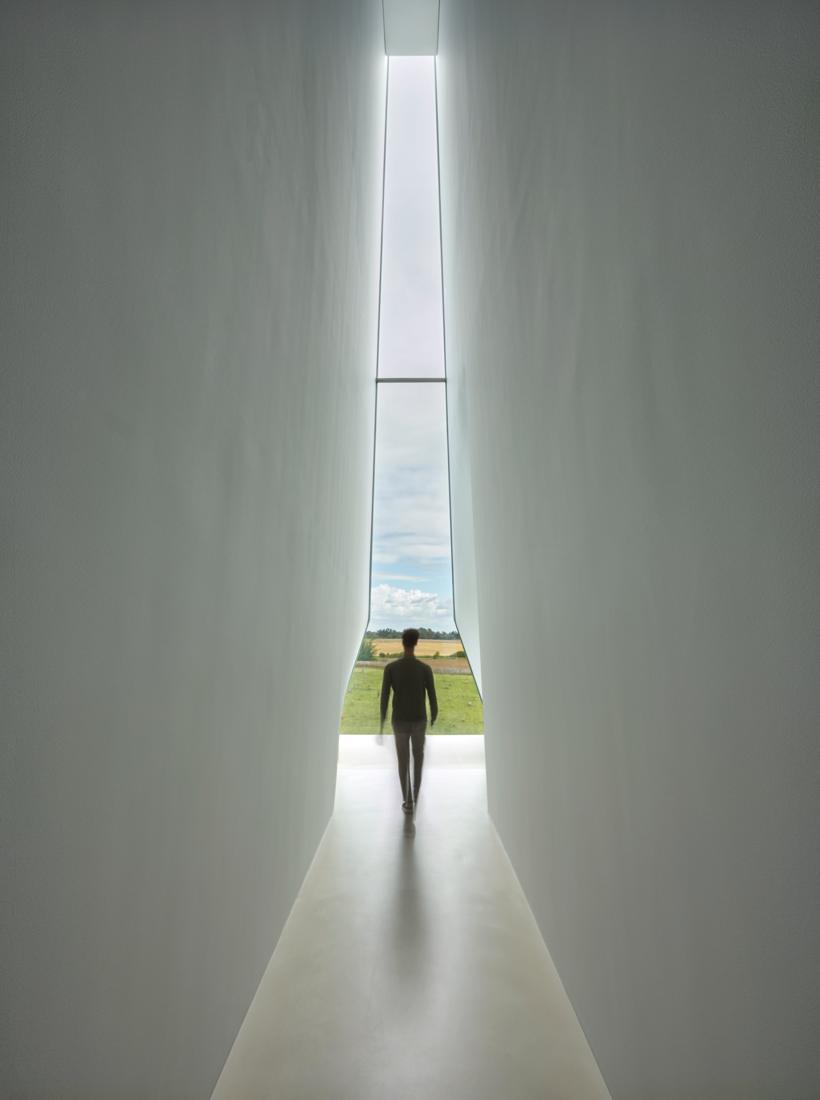 A figure walks down a narrow white corridor toward a window overlooking the fields