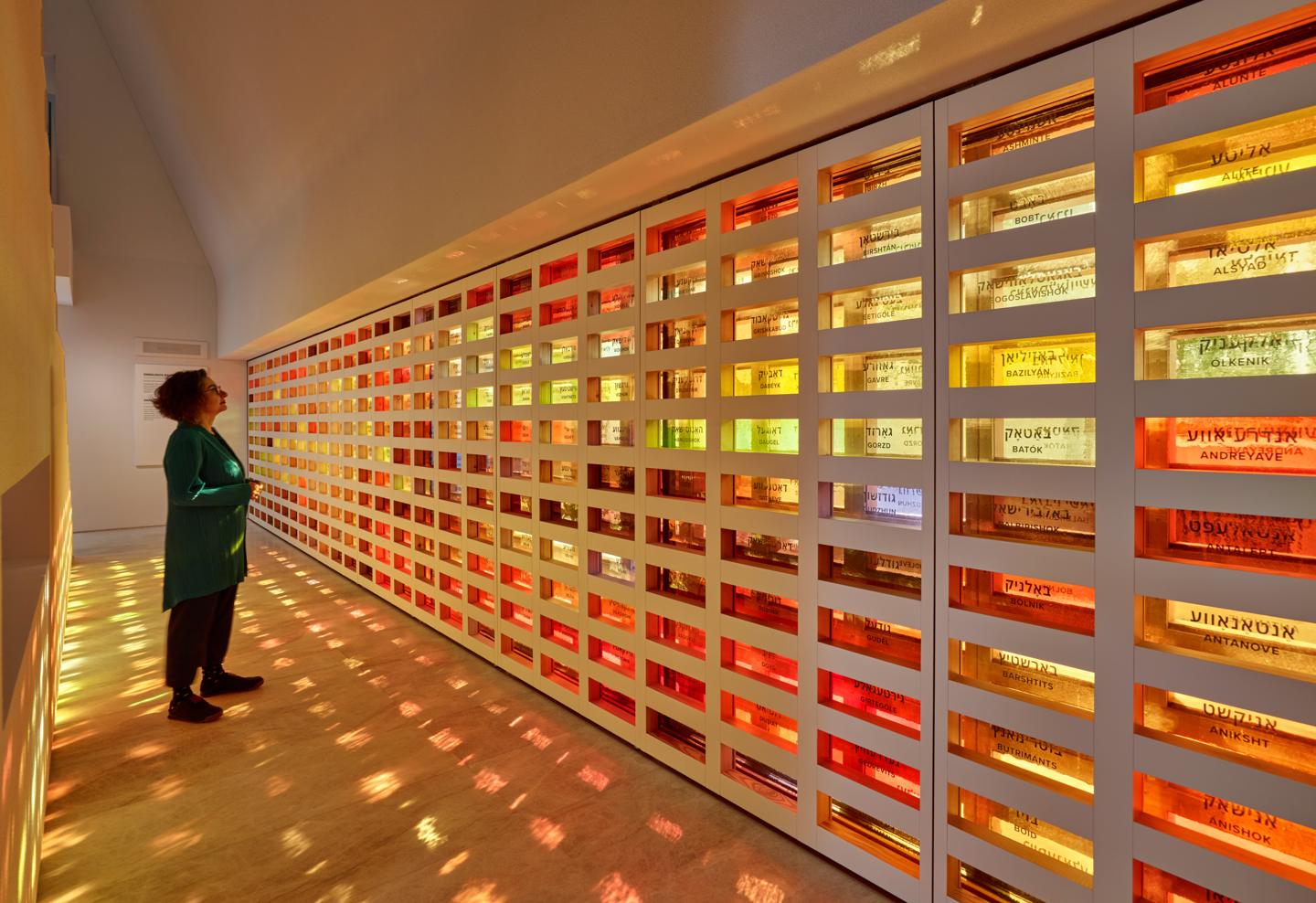 A visitor before a wall of coloured glass panels bearing the names of Lithuanian Jewish communities