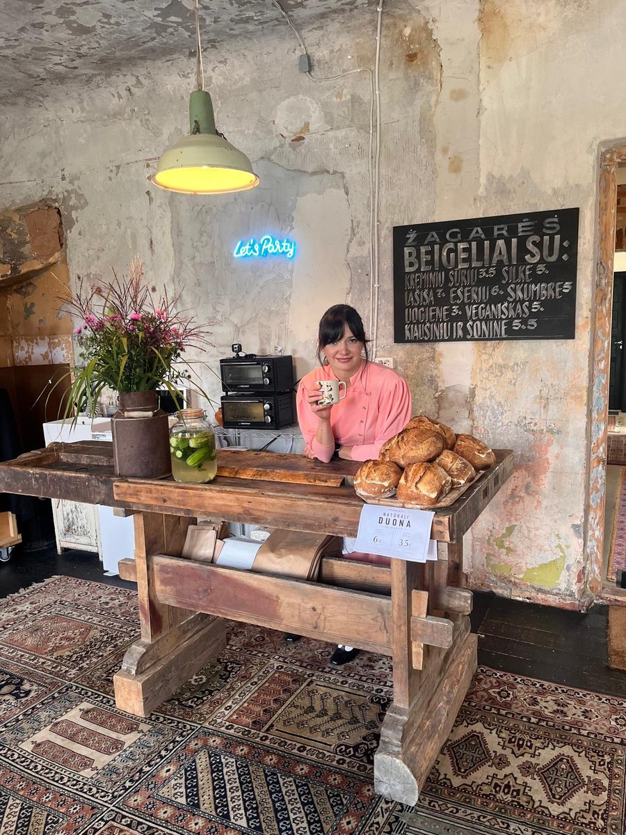 Woman at a wooden counter with baked goods and a chalkboard sign for Žagarės Beigeliai